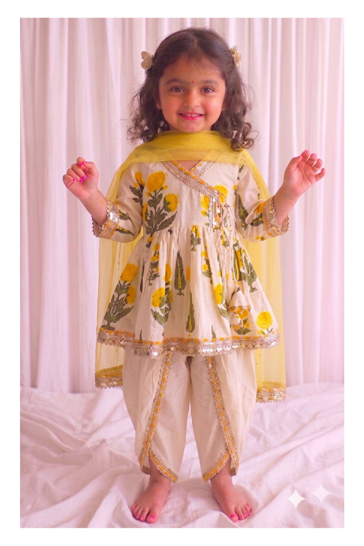 A young girl stands barefoot in front of a white curtain backdrop, wearing a cream-colored traditional outfit with yellow floral prints, gold trim, and a yellow dupatta.