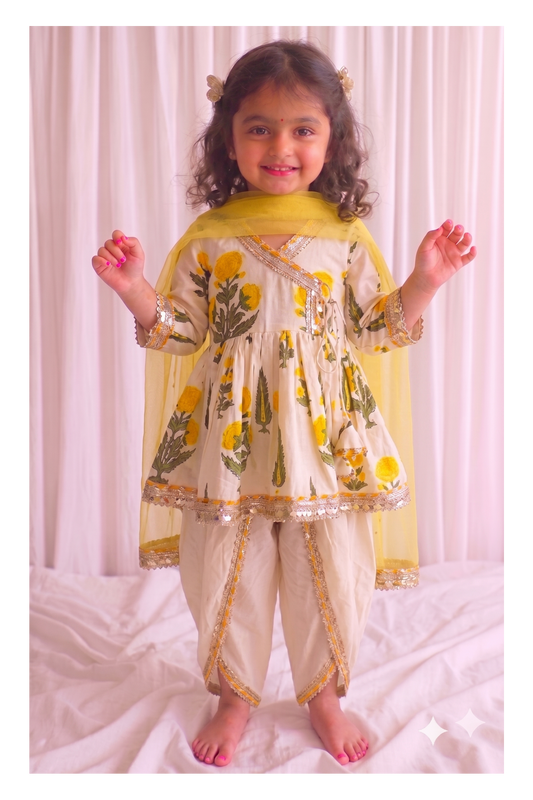 A young girl stands barefoot in front of a white curtain backdrop, wearing a cream-colored traditional outfit with yellow floral prints, gold trim, and a yellow dupatta.