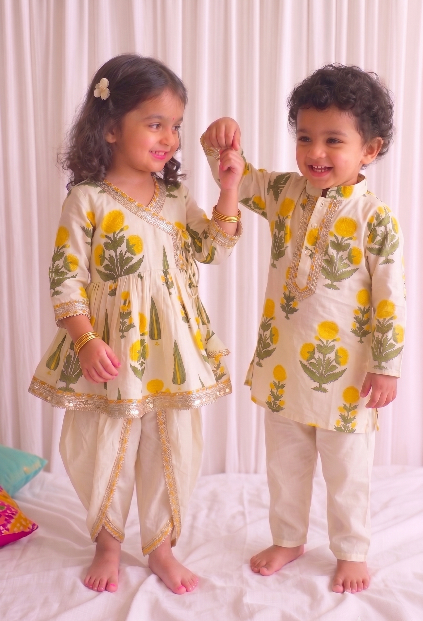Two young children modeling matching cream-colored traditional Indian outfits with yellow floral prints and gold borders, standing on a white bed against a white curtain backdrop.