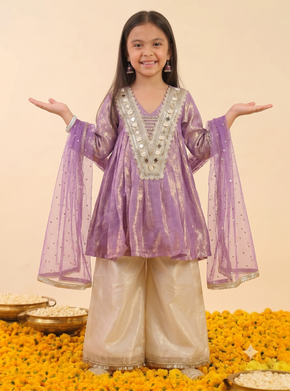 A young girl stands with arms outstretched wearing a lavender purple traditional kurta set with silver mirror work on the neckline and wide-leg metallic beige pants. She is posed on a bed of yellow marigold flowers with brass bowls in the background against a solid cream backdrop.