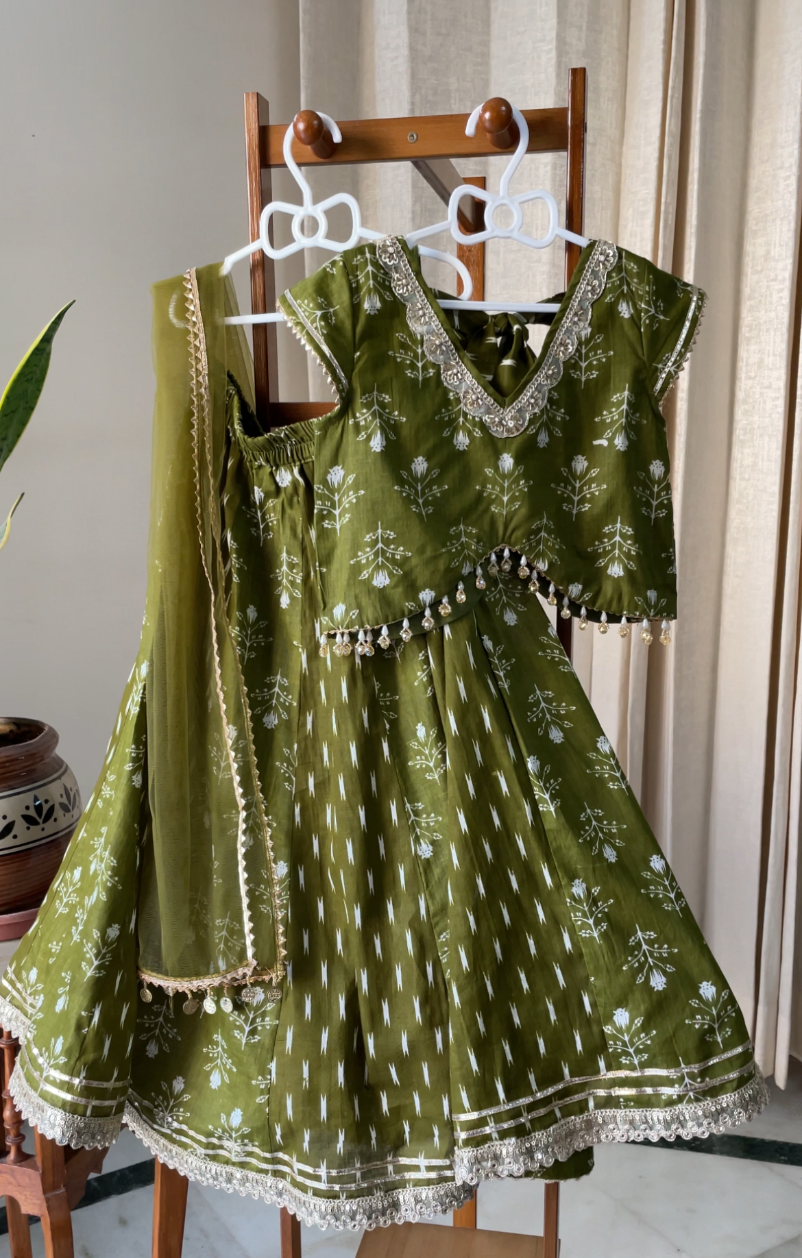 An olive green traditional Indian dress set with white floral prints and gold embroidery, displayed on a wooden hanger stand. The set includes a blouse, flared skirt, and dupatta.