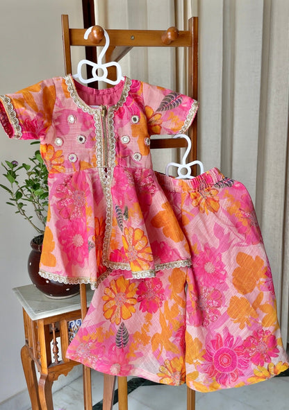 A two-piece baby girl's floral print outfit consisting of a short-sleeve peplum top and matching sharara, displayed on a wooden stand. The outfit features a vibrant pink, orange, and yellow floral pattern with silver mirror embroidery along the neckline.