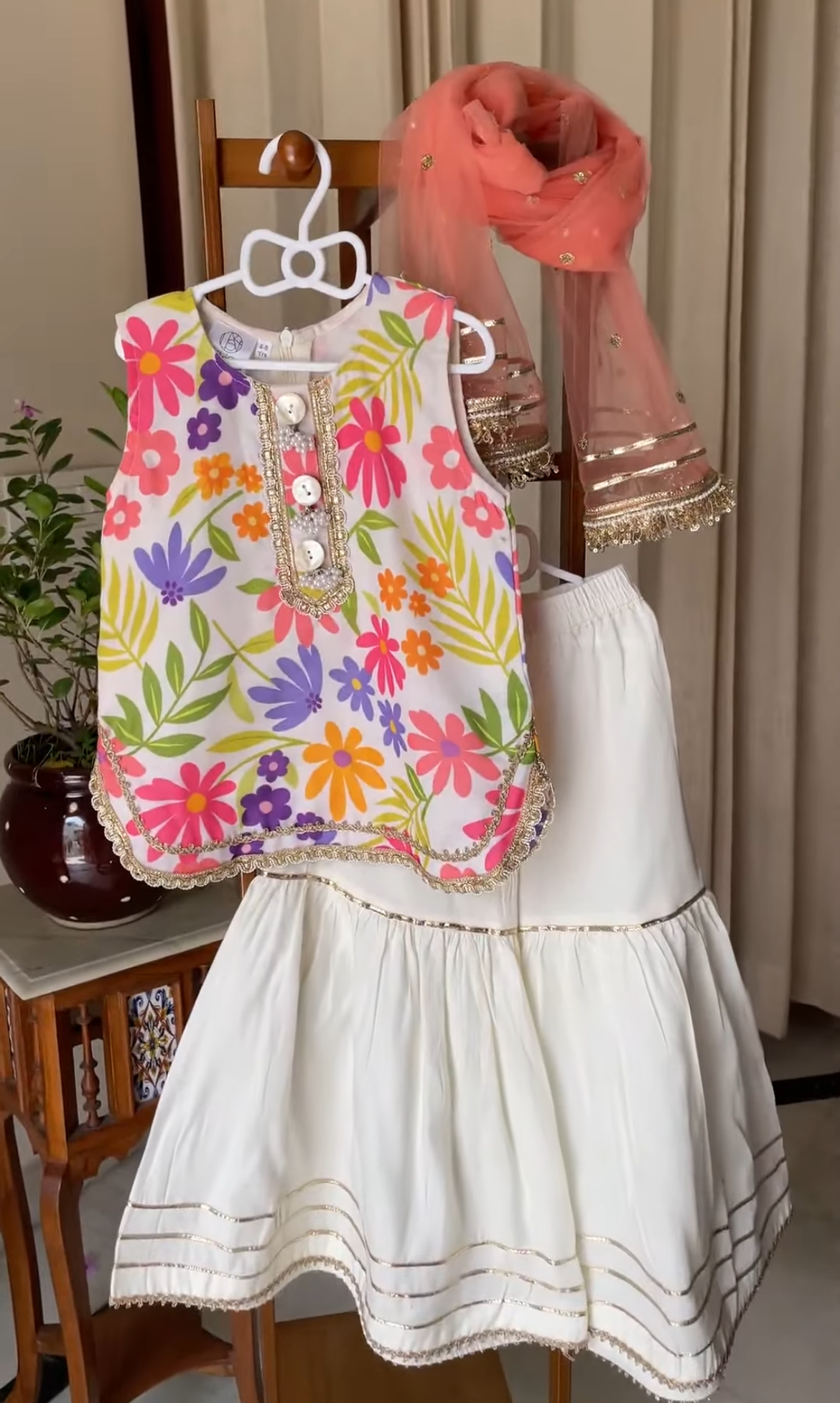 A children's outfit displayed on a hanger in an indoor setting, featuring a sleeveless white top with a colorful floral print in pink, orange, yellow, green, and purple, paired with a white sharara and a peach dupatta.
